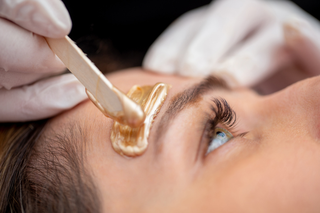 Beautician Applying Wax with Wooden Spatula Above Woman's Eyebrow - stock photo